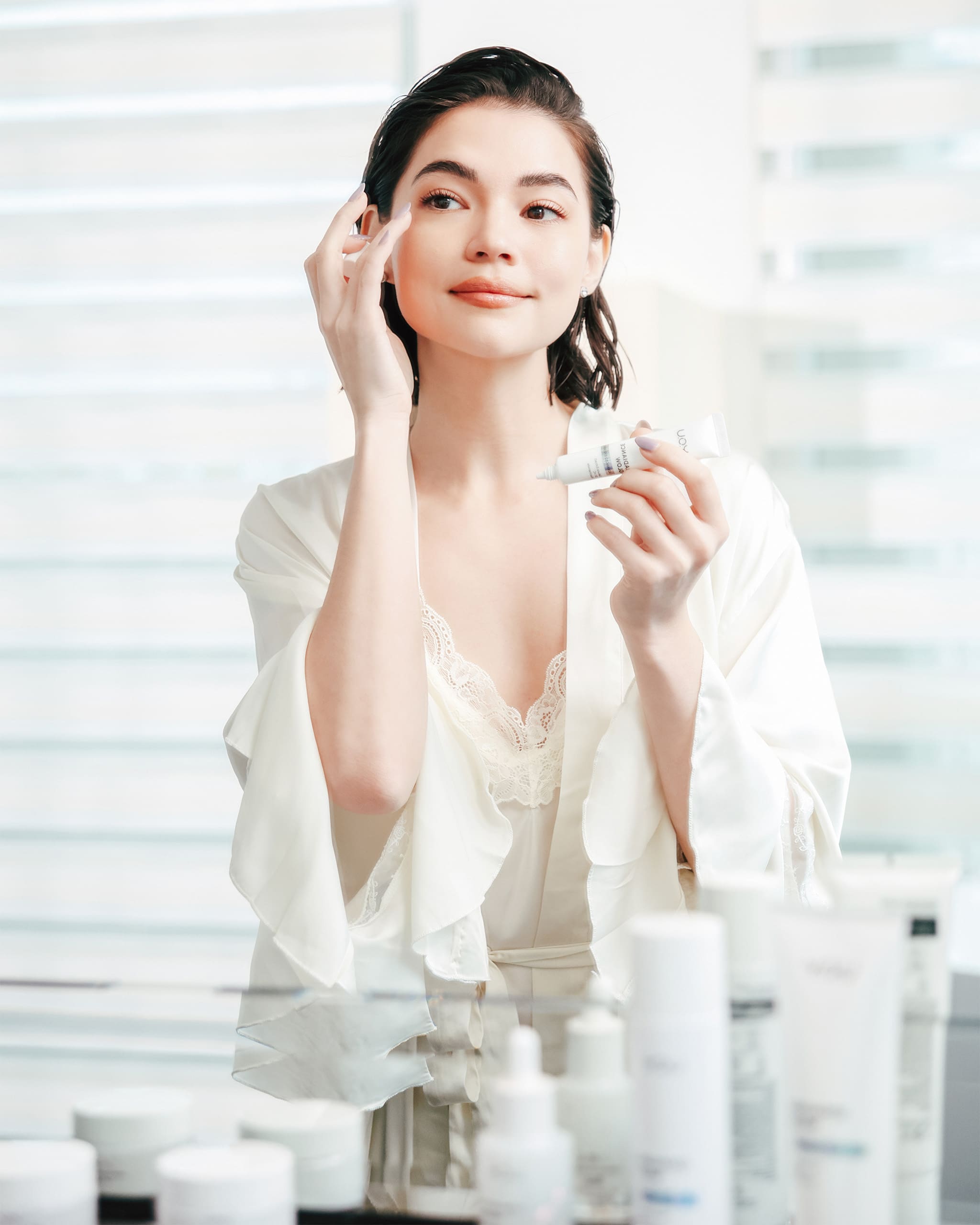 Rhian Ramos looks at her reflection in a mirror, applying Y.O.U Beauty Radiance Glow skincare for a radiant complexion.